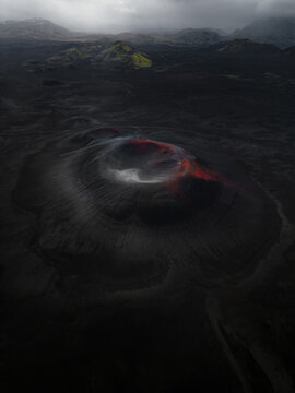 Aerial view of a volcanic crater with red mineral deposits surrounded by dark lava fields and mossy hills in Iceland.