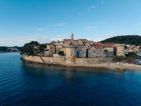 Aerial view of ancient stone buildings with red-tiled roofs meet the turquoise sea, framed by lush greenery, a timeless scene of coastal beauty, Korcula, Dubrovnik-Neretva County, Croatia.