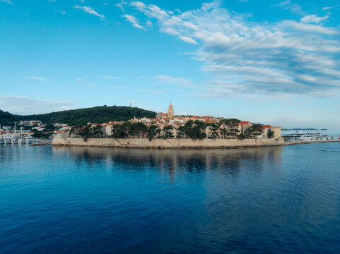 Aerial view of the medieval walled city shimmering in the turquoise waters, framed by verdant hills under a sky streaked with wispy clouds, Korcula, Dubrovnik-Neretva County, Croatia.