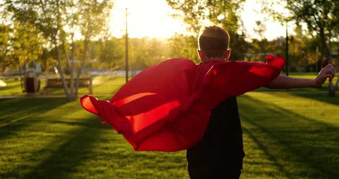 Superhero boy runs across green park at sunset. Superhero cape spreads wide in golden light. Child imagines hero future and dreams big outdoors. Children's freedom, courage, playful childhood.