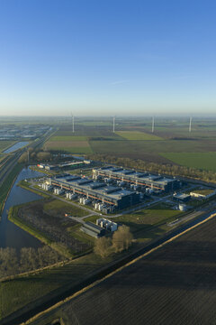 Aerial view of a modern industrial complex nestled amidst patchwork fields and waterways, with wind turbines standing tall on the horizon, Middenmeer, North Holland, Netherlands.