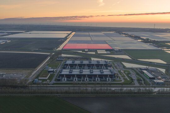 Aerial view of a sprawling industrial complex casting long shadows as the sun dips low, painting the sky with fiery hues, Middenmeer, North Holland, Netherlands.