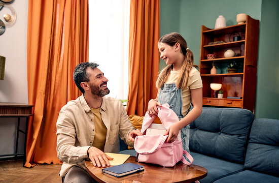 Back to school preparation.Father and daughter are getting ready for the first day of school. Father helps his child pack a pink backpack with books in a colorful living room.