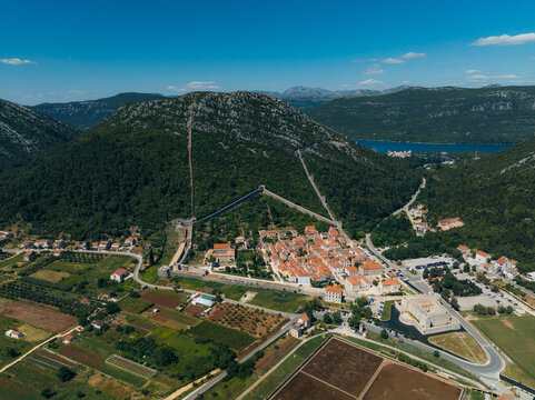 Aerial view of the ancient Walls of Ston snaking through lush green hillsides, contrasting with the terracotta rooftops of the town below, Ston, Dubrovnik-Neretva County, Croatia.
