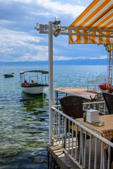 Fototapeta premium A sunny restaurant terrace with striped awning and outdoor seating offers views of Lake Ohrid, where boats gently float on the blue-green water.