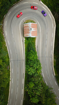 Aerial view of vibrant cars navigating a hairpin turn around a quaint building amidst lush greenery, Gex, Auvergne-Rhone-Alpes, France.