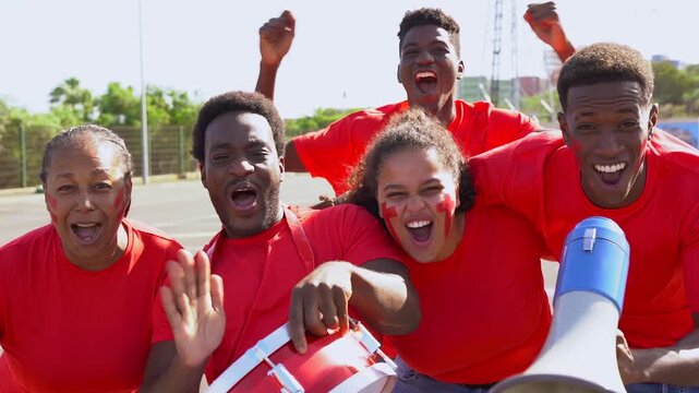 Group of african football supporter fans watching soccer match event at stadium - Multi generational people with red t-shirts having fun together on sport world championship event