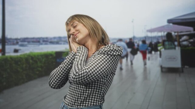 Woman resting hands to cheek in a houndstooth sweater on a waterfront street promenade, eyes closed and smiling; serenity.