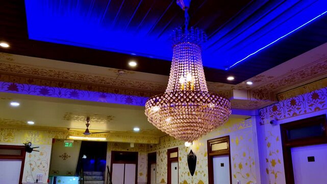 An ornate indoor hall in Jaipur, India, with a large crystal chandelier hanging from a wooden ceiling, blue LED lighting around borders, and floral patterned walls giving a royal interior look.