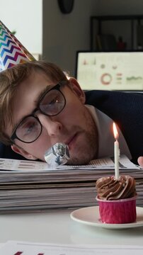 Vertical shot of fatigued gen Z male employee with colourful hat using party blower and watching white candle burning on chocolate cupcake with his head on pile of documents