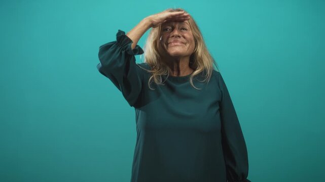 Senior blonde woman shields her eyes with hand and smiles while looking upward in studio wearing teal dress; calm contentment.
