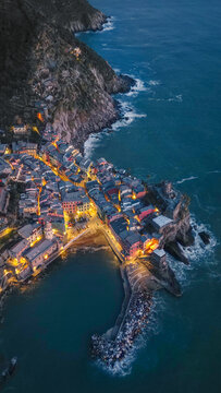 Aerial view of the illuminated village of Vernazza with its historic harbor and colorful houses nestled along the rugged coastline at twilight in Vernazza, Liguria, Italy.