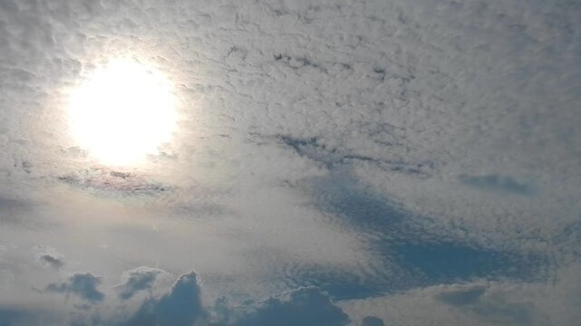 Slowly moving natural clouds texture Altocumulus Duplicatus in blue sky with sun and colorful pearly glow around sunlight. Topics: cloud iridescence, irisation, optical phenomenon, meteorology