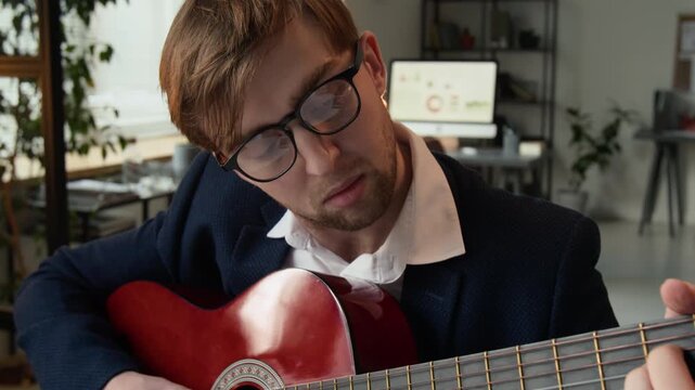 Tilt up view of formally dressed gen Z businessman with glasses strumming chords using wooden guitar in office setting