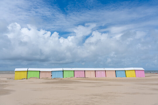 View of pastel-colored beach huts line the sandy shore under a vast, cloud-streaked sky, creating a tranquil coastal scene, Berck-Plage, Hauts-de-France, France.