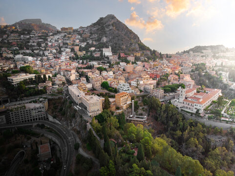 Aerial view of the historic hillside town with traditional rooftops, lush greenery, and a winding road under a golden sunset sky Taormina, Sicily, Italy.