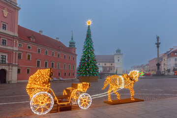 Obraz premium Christmas tree and illuminated horse carriage decoration in Castle Square, Warsaw, Poland, at dawn in winter fog.
