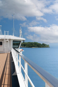 View of the white bridge wing and railing of a ship approaching the lush tropical coastline under a blue sky with white clouds Iles du Salut, Guyana.