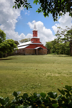 View of the historic red and white church with a stone base surrounded by lush green trees and a grassy field under a cloudy sky in Iles du Salut, Guyana.