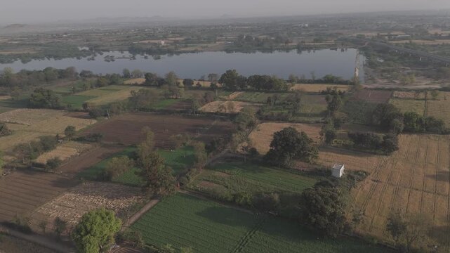 Aerial view of agricultural fields in Bhilwara, Rajasthan during spring harvest, showing half-harvested wheat, crop bundles forming abstract patterns, with the Kothari River flowing through farmland.