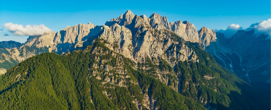 Aerial view of jagged mountains rise majestically above lush green forests under a clear blue sky, Bohinjsko jezero, Kranjska Gora, Slovenia.