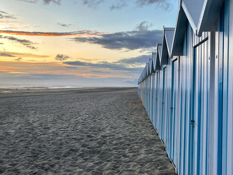 View of beach huts stand in neat rows along a sandy beach under a pastel sky at dusk, their blue and white stripes mirroring the sea, Fort-Mahon-Plage, Hauts-de-France, France.