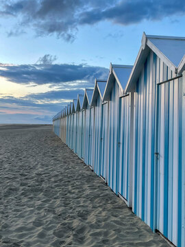 View of blue and white striped beach huts stand in orderly rows along the sandy shore under a soft, cloudy sky, Fort-Mahon-Plage, Hauts-de-France, France.