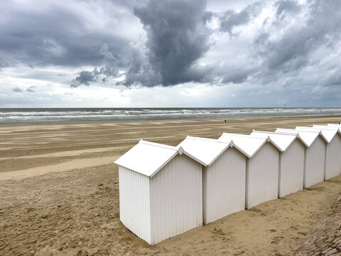 View of pristine white beach huts stand in orderly rows against the backdrop of a sandy beach and a brooding, stormy sky, Fort-Mahon-Plage, France.