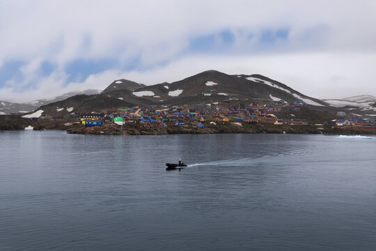 View of colorful houses in a remote coastal village at the base of rocky hills with a small boat on the water in Ittoqqortoormiit, Sermersooq Municipality, Greenland.