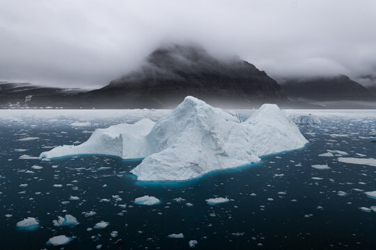 View of a massive white iceberg floating in dark blue water with misty mountains and low clouds in Greenland.