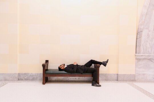 Side view of a businessman in a suit and sunglasses lying down on a wooden bench in a lobby