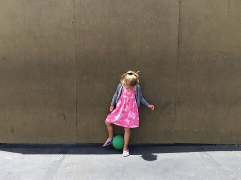 Young girl in a summer dress and flip flops standing in front of a wall playing with a ball