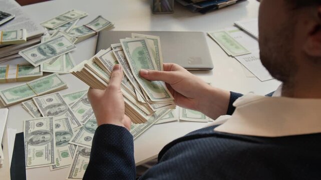 Over the shoulder shot of formally dressed young man counting freshly printed dollar banknotes sitting at white office desk with laptop, business papers and money on it