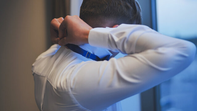 Groom getting ready for wedding, adjusting his blue bow tie in front of a window. Back view.