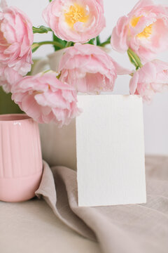 Close-up of a blank card leaning against a white jug with a bouquet of Pink peony tulips (double tulips), a ribbed mug and a linen napkin