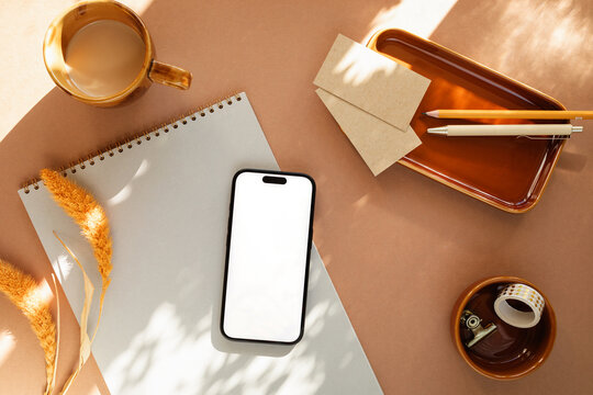 Overhead view of a mobile phone with a blank screen on a notepad with a cup of white coffee, ceramic tray, blank business cards, pens, a scented candle, napkin and dried pampas grass