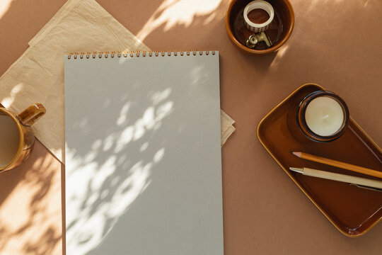 Overhead view of a blank notebook, cup of white coffee, scented block candle, pens, washi tape and a folded napkin on a table in sunlight