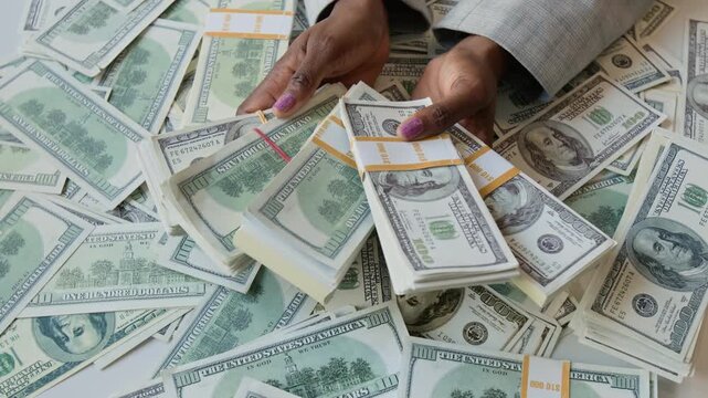 Close-up shot of hands of anonymous Black woman holding five stacks of dollar bills, with banknotes scattered across white table