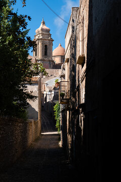 View of a narrow cobblestone alleyway leading towards a historic church tower and dome under a clear blue sky in Segesta, Sicilia, Italy.