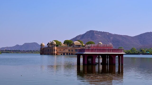 Video view of Jal Mahal in Man Sagar Lake Jaipur with red sandstone pavilion chhatri structure in foreground, Aravalli hills in background and lakeside Jal Mahal viewpoint area visible on right side.