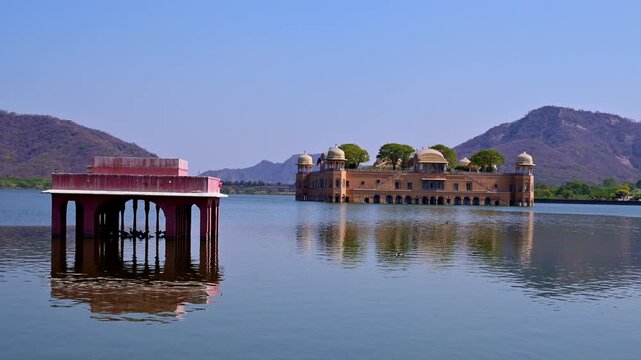 Wide video shot of Jal Mahal in Man Sagar Lake Jaipur with red chhatri pavilion in foreground, reflections in water, flying bird over lake and Aravalli hills behind palace.
