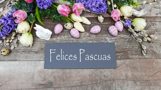 D&eacute;coration de P&acirc;ques avec des fleurs violettes et blanches, des &oelig;ufs roses, une figurine de lapin blanc et une pancarte portant l'inscription espagnole &laquo; Felices Pascuas &raquo; sur un fond en bois.