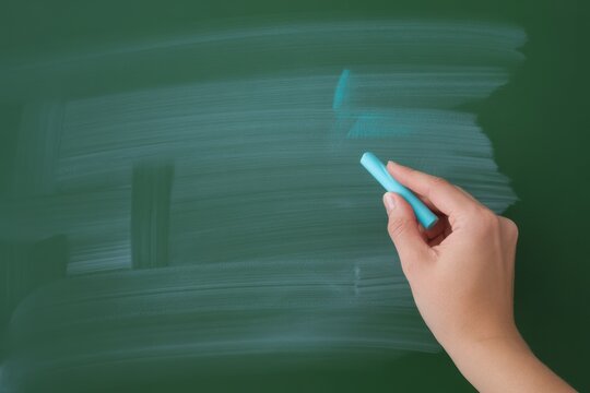 A hand holding blue chalk erases writing on a green chalkboard.