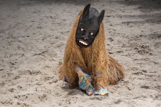 View of a person wearing a traditional mask and raffia costume sitting on the sand in Freetown, Western Area, Sierra Leone.