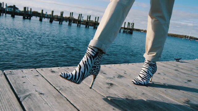 Unrecognized woman in light jeans and high-heeled boots with zebra print walks by the wooden pier. Stylish footwear and fashion.