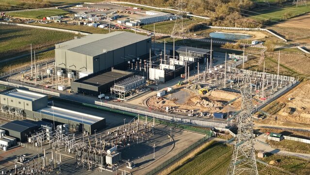 Aerial view of the vast Bramford Substation, a symphony of steel and power infrastructure against a backdrop of golden fields and azure skies, Ipswich, United Kingdom.