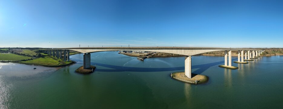 Aerial view of a concrete bridge soaring over the rippling water, casting shadows on the green water under a clear blue sky, Orwell Bridge, Ipswich, United Kingdom.