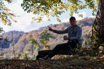 Fototapeta premium Male relaxing with beverage in fall forest landscape. Man in light blue jacket sits on wooden log sipping from cup amid yellow autumn leaves and mountainous backdrop.