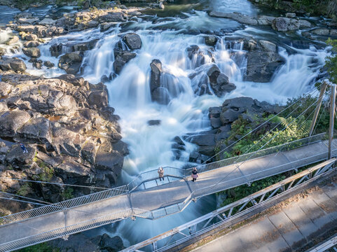 Aerial view of a vibrant waterfall cascading over rugged rocks, embraced by lush greenery and a pedestrian bridge, Lower Kaimai, New Zealand.