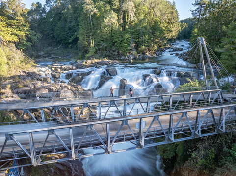 Aerial view of cascading waters plunge over rocky ledges, framed by verdant forests and a sturdy bridge where two figures stand, Lower Kaimai, New Zealand.